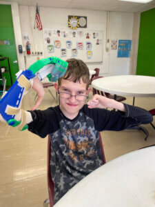 A young boy smiling while flexing both biceps with attached fabricated upper limb device on his right arm